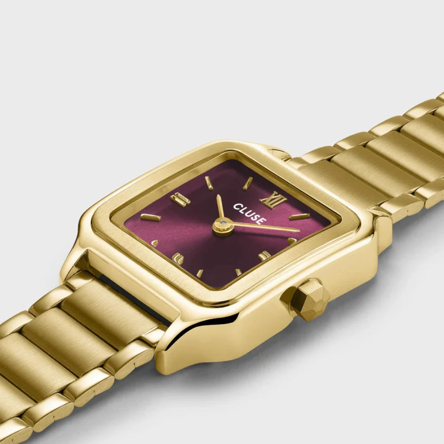 a side shot of a  gold coloured cluse watch with a maroon face laid on a white background